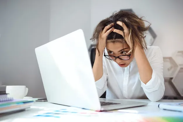 A woman in a white shirt and glasses holds her head in her hands, looking frustrated or stressed, in front of a laptop on a desk cluttered with papers and supplies.